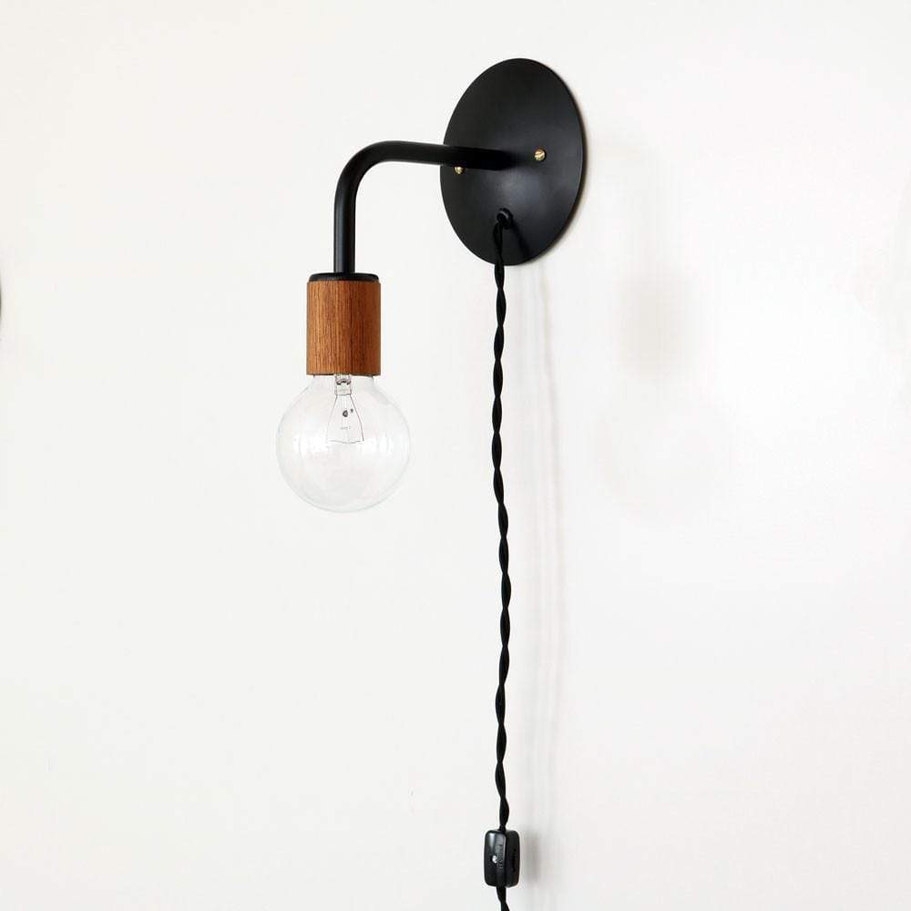 A minimalist wall-mounted light fixture featuring a black circular mount with a black curved arm leading to a wooden socket, from which hangs a clear, round light bulb.