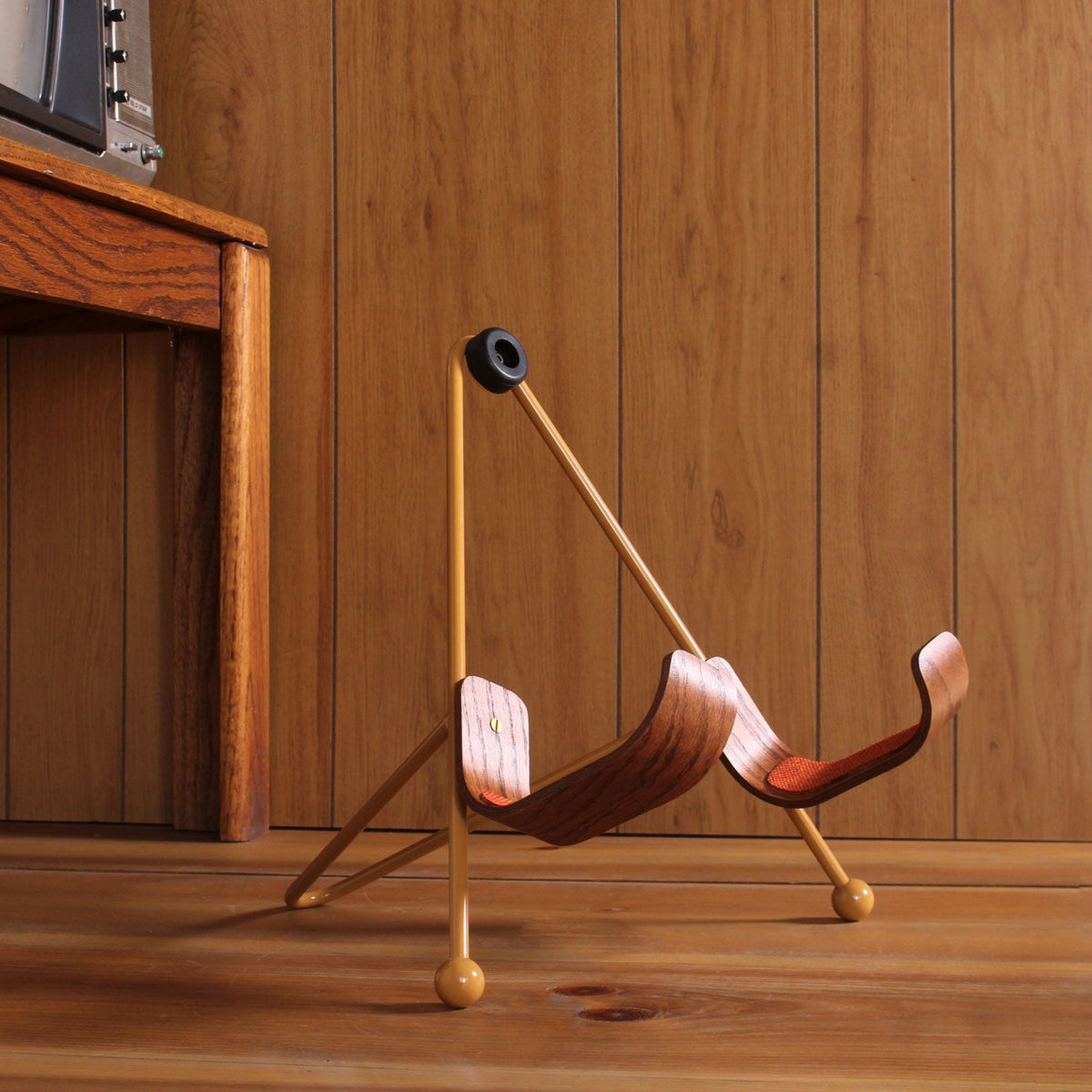 VHS Edition Guitar Stand with cream base and stained oak rests with houndstooth tweed, shown next to a vintage CRT TV and wood paneling.