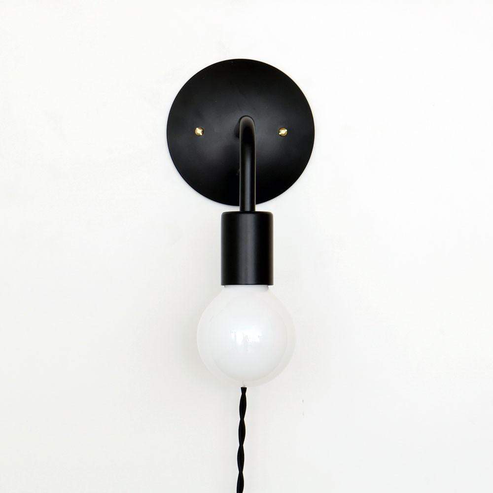A minimalist wall-mounted light fixture featuring a black circular mount with a black curved arm leading to a wooden socket, from which hangs a clear, round light bulb.